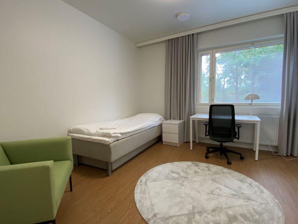 Ikkunallinen huone, jossa on sänky ja yöpöytä, nojatuoli, työpöytä ja -tuoli, ikkunoissa verhot ja lattialla matto.