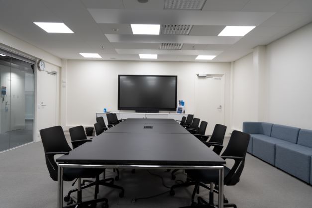 Kokoushuone 12 henkilölle; pöytä jonka äärellä 12 tuolia, seinällä näyttö, sivulla sohva. Toisella sivulla ikkunaseinä.