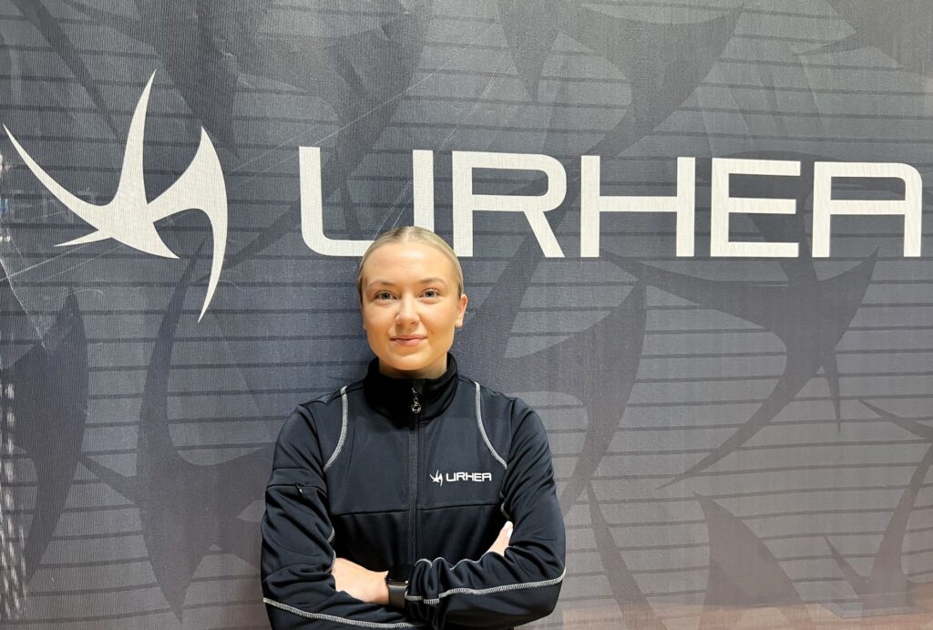 Veera Törmä seisoo Urhea-logon edessä, tummansininen tausta ja logo valkoisella