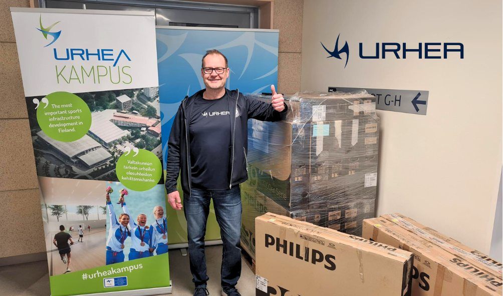 Mika Saari seisoo televisiolaatikoihin nojaten ja näyttää peukkua, taustalla Urhea-kyltti